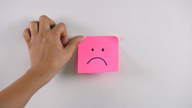 Woman's hand sticking note with a sad face emoticon over a white background. Closeup shot of a pink color sticky note stuck to the white wall with a sad face emoji - negative emotions concept