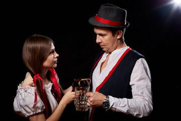 Girl pouring beer from a keg into a mug of guy in Octoberfest. Couple in nationa ethnic dress having fun with alcohol. Girlfriend and boyfriend posing in studio with black background in Oktoberfest.