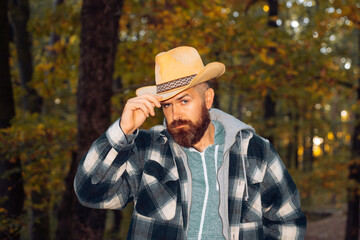 Brutal brunette bearded man in warm hat in the woods on a background of trees. Stylish hipster man in the autumnal forest. Macho man at autumnal nature.