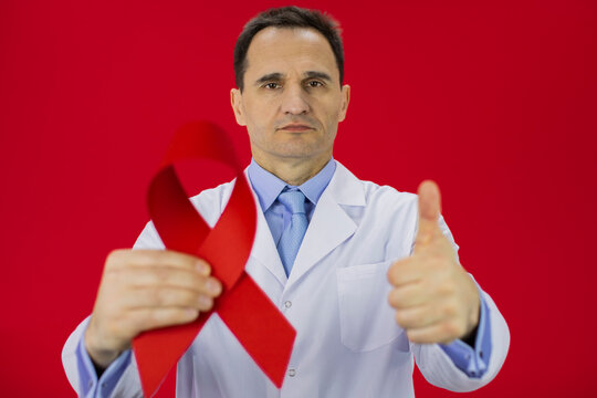 Handsome serious male senior age doctor in white lab coat with red ribbon in hand shows like sign with thumb up on red background, focus on doctor. Symbol of HiV Aids struggle, awareness, medical sign - Powered by Adobe