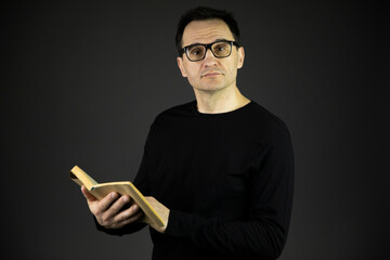 Attractive intelligent man in casual black wear and glasses holding classic book in hands and thoughtfully looks at camera isolated on dark background. Reading, education, relaxing, writer or author