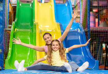 Smiling mother and her daughter riding slide together at indoor kids playground