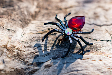 An extravagant women's brooch in the form of a black spider with rhinestones and a red stone, shimmering in the sunlight under the open sky on a wooden surface. Red forest ants attack the spider