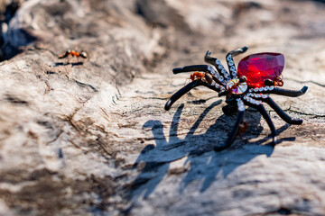 An extravagant women's brooch in the form of a black spider with rhinestones and a red stone, shimmering in the sunlight under the open sky on a wooden surface. Red forest ants attack the spider