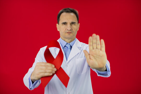 Handsome serious male senior age doctor in white lab coat with red ribbon in hand shows stop sign isolated on red background Symbol of HiV Aids struggle, awareness, medical sign, world aids day - Powered by Adobe