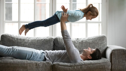 Smiling young father lying on couch in living room hold in arms imitate plane with little preschooler daughter, happy Caucasian dad have fun playing on sofa with overjoyed small girl child at home