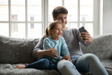 Smiling young Caucasian father and small preschooler daughter sit on sofa at home make self-portrait picture on cellphone, happy dad and little girl child take selfie on smartphone gadget together