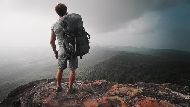 Backpacker with valley view. Hiker man stands at the edge of the mountain taking backpack to shoulder