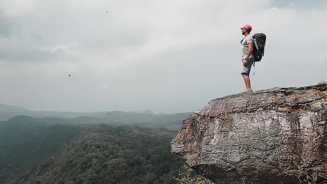 Backpacker with valley view. Hiker man stands at the edge of the mountain taking backpack to shoulder and leaves