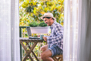 Attractive young man laughs happily while talking to his laptop on a tropical balcony. He holds his coffee in one hand and wears a hat.