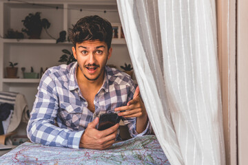 Attractive young man makes a surprised gesture looking at the camera with the mobile phone in his hand. He's behind the curtain with a map in front of him.