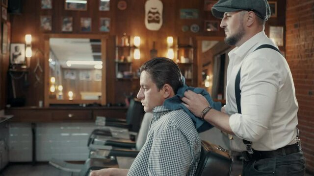 Stylishly dressed Barber dancing in a barbershop.
Brutal Barber with a beard wipes the hair of the client before cutting. Dancing Barber.