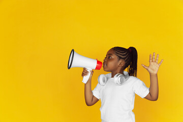 Shouting with megaphone, listen to music. Little african-american girl's portrait on yellow studio...