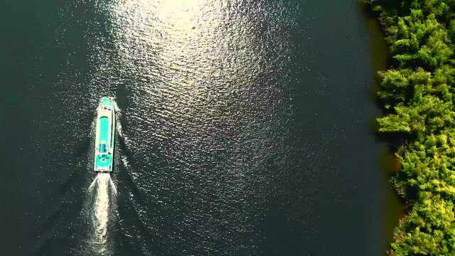 Vertical rone fly over a passenger boat on a river next to beautiful green trees and coastline. China.