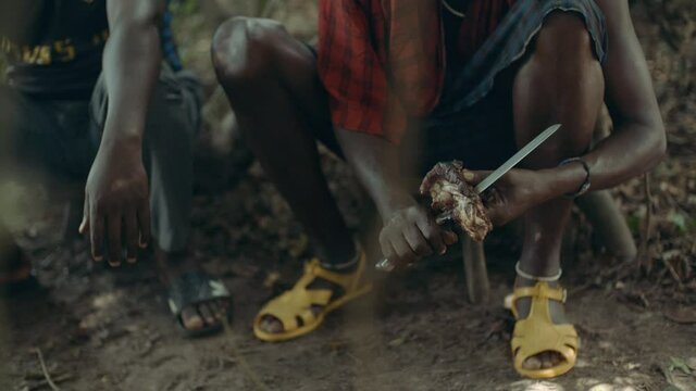 An African sitting in a squat and wearing traditional Maasai clothes is cutting a piece of meat with a knive. We can not see their faces but the dancing sunlight on the ground.