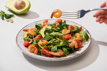 partial view of woman eating fresh green salad with cherry tomatoes, shrimps and avocado on white background