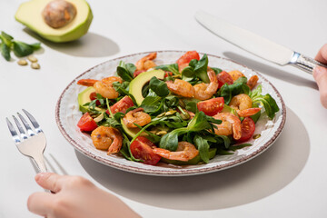 partial view of woman eating fresh green salad with cherry tomatoes, shrimps and avocado on white background