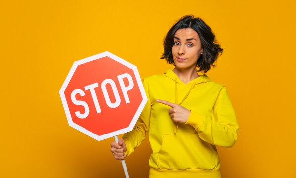 Persistent requirement. Half-length photo of confident and persistent young girl, dressed in a yellow hoodie, earnestly pointing on "stop" sign with her finger.