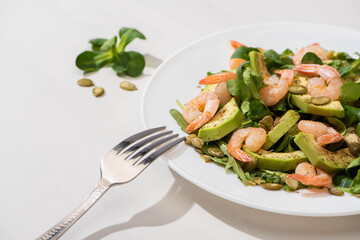 selective focus of fresh green salad with shrimps and avocado on plate near fork on white background