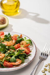 selective focus of fresh green salad with shrimps and avocado on plate near fork and ingredients on white background