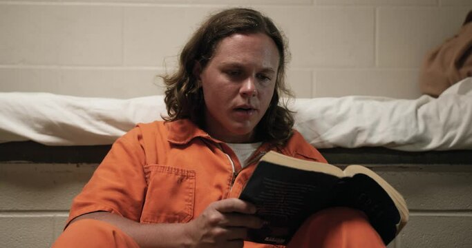 A man sits in his prison cell reading a bible while wearing an orange jumpsuit