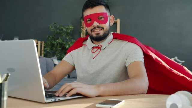 Slow motion portrait of Middle Eastern man in superman cape and mask at computer desk at home smiling and looking at camera. People and lifestyle concept.