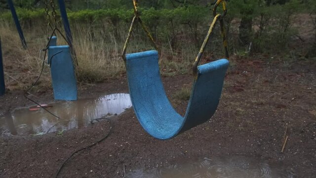 Old broken blue and yellow swing set swaying in a children's playground close up