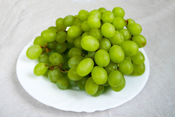 Grona of white seedless grapes in a white plate on a beige linen tablecloth . raw fruit on a plate