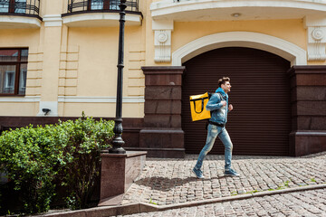 Side view of smiling courier with thermo backpack walking on paving stone walkway near buildings