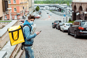 Side view of courier in medical mask with thermo backpack using smartphone on urban street