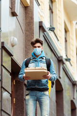 Fototapeta na wymiar Courier in medical mask looking at camera while holding pizza boxes on urban street
