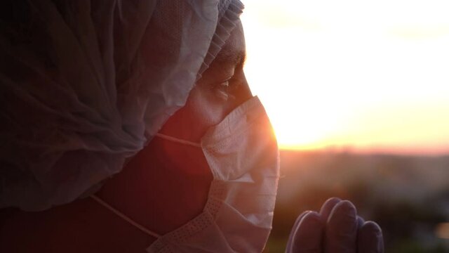 Doctor in a protective mask and gloves prays to God at sunset, close-up