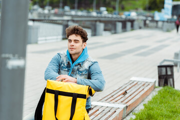 Selective focus of courier sitting near thermal bag on bench on urban street