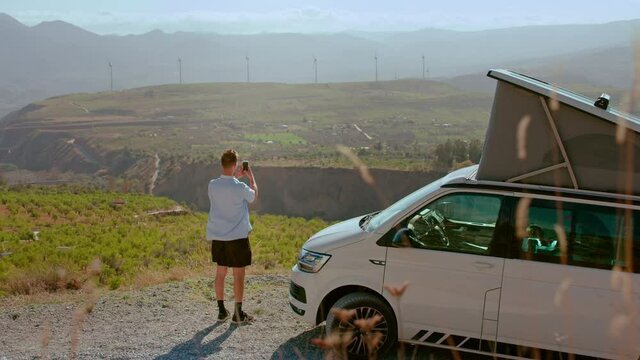 Tourist traveler man makes photos on smartphone of epic landscape. Man stand next to camping RV van up a mountain in beautiful scenery. Road trip adventure and camp vibes for staycation