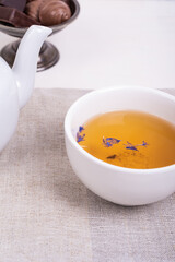 Cup of green tea with flower petals on a linen napkin and candies in a vintage metal vase on a white wooden table, close-up