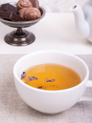 Cup of green tea with flower petals on a linen napkin and candies in a vintage metal vase on a white wooden table, close-up