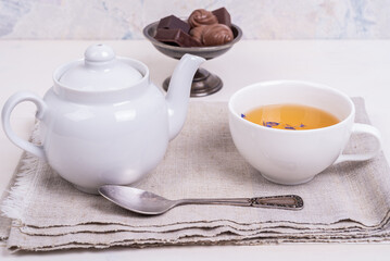 Teapot, cup of green tea with flower petals and candies in a vintage metal vase on a white wooden table, close-up