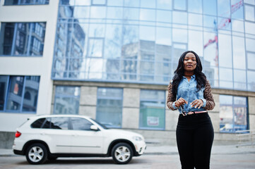 Hipster african american girl wearing jeans shirt with leopard sleeves posing at street against modern office building with blue windows and white suv car.