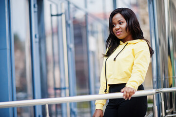 Hipster african american girl wearing yellow hoodie posing at street against office building with blue windows.