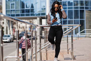 Hipster african american girl wearing sunglasses, jeans shirt with leopard sleeves, hold mobile phone, posing at street against modern office building. - obrazy, fototapety, plakaty