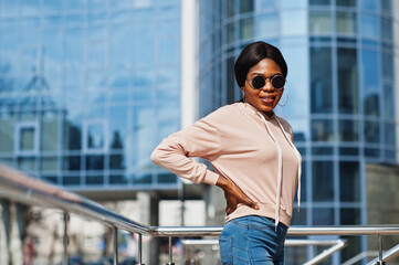 Hipster african american girl wearing pink hoodie,sunglasses and jeans posing at street against office building with blue windows.