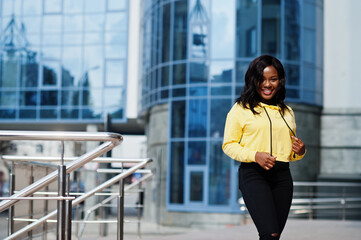 Hipster african american girl wearing yellow hoodie posing at street against office building with blue windows.