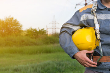 A young electrical engineer, with a protective helmet in his hands, against the background of a power line. Close - up of the worker