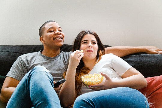 Interracial couple sitting on the sofa watching a movie and eating popcorn