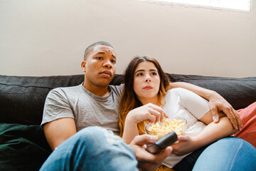 Interracial couple sitting on the sofa watching a movie and eating popcorn