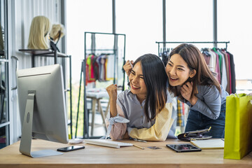 Two young Asian girls shop owners at work desk inside a clothing store looking and pointing at computer monitor with happy cheerful wow surprised expression.