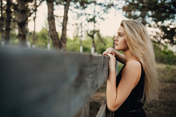 Young blonde attractive woman with long hair and blue eyes, standing by a wooden fence, outdoors, looking to side.