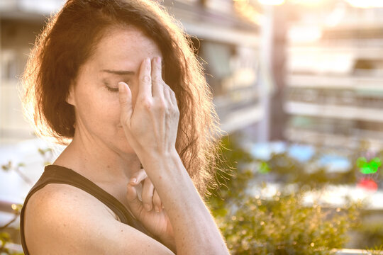 Portrait of a mature woman with hear hand on forehead and closed eyes, worry, headache, facing problems, urban background.