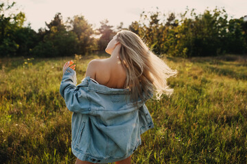 Young attractive woman with long blonde hair, standing in a field with wind in hair, outdoors, wearing a denim jacket.