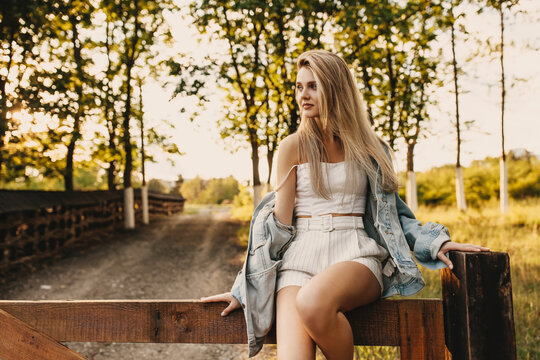Young happy woman with long blonde hair, outdoors, wearing a denim jacket and a crop top, sitting on a wooden fence.